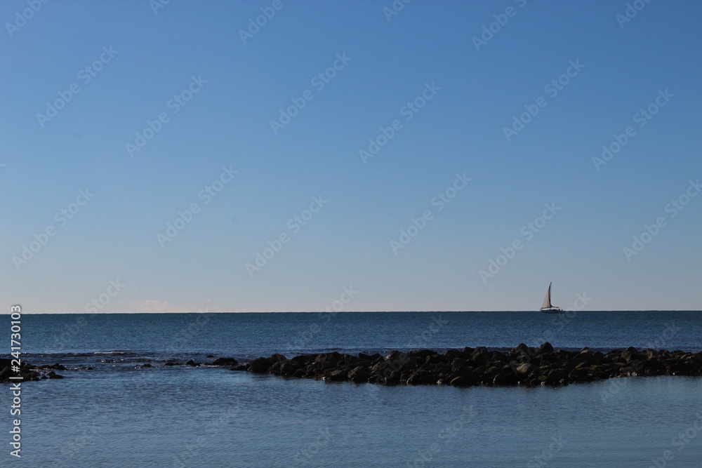 barca a vela in mare all'orizzonte con scogli e cielo azzurro