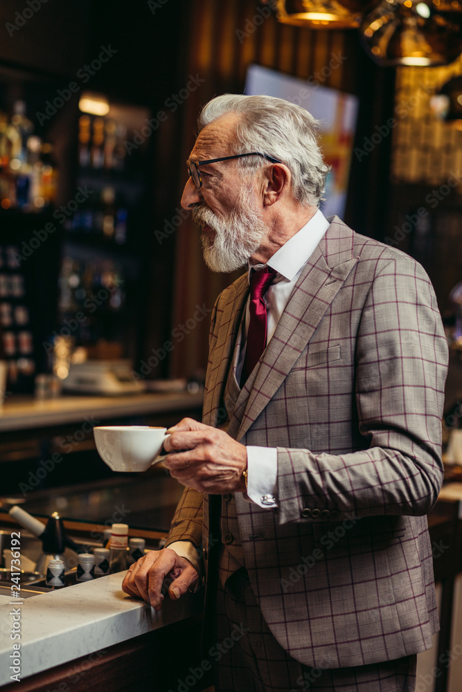 Senior businessman in rich formal three piece suit drinking coffee standing near bar counter at dark restaurant interior and looking at camera Senior businessman in rich formal three piece suit drinking coffee standing near bar counter at dark restaurant interior and looking at camera