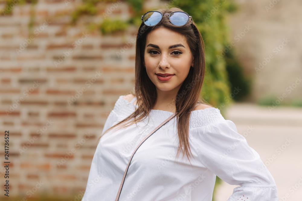 Portrait of happy female in sunglasses. Girl is dressed in a white shirt is standing near a wall overgrown with ivy Portrait of happy female in sunglasses. Girl is dressed in a white shirt is standing near a wall overgrown with ivy