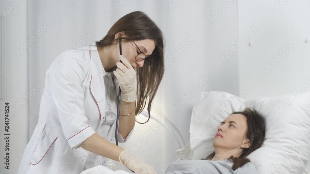The doctor conducts a routine examination of a patient in a hospital ward. Woman listening the patient with stethoscope in bed in a hospital ward. The doctor conducts a routine examination of a patient in a hospital ward. Woman listening the patient with stethoscope in bed in a hospital ward.