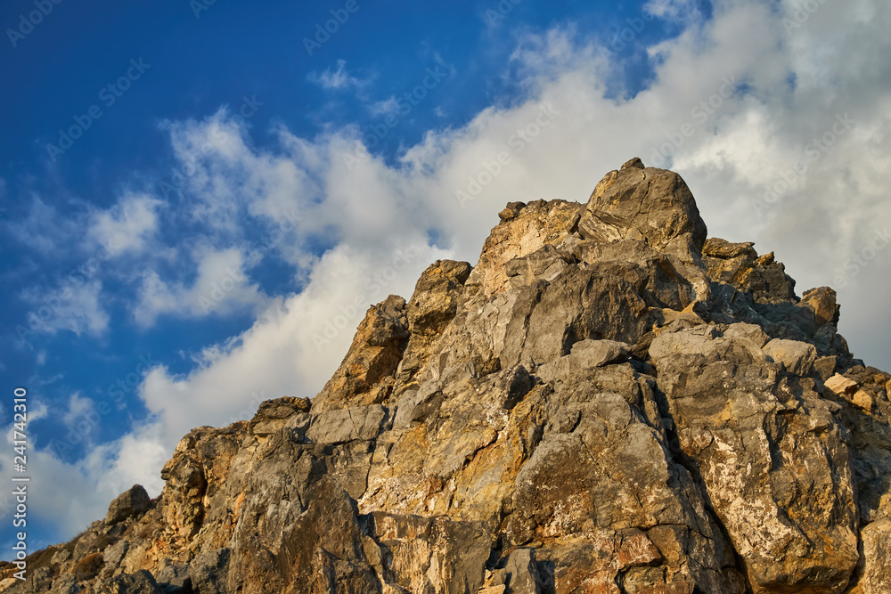 Coastal rocky peak of a cliff with white clouds on a blue sky in summer in Greece.