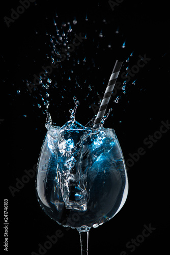 Ice cube falling into a glass with a cocktail and a striped tube on a black background with blue light.