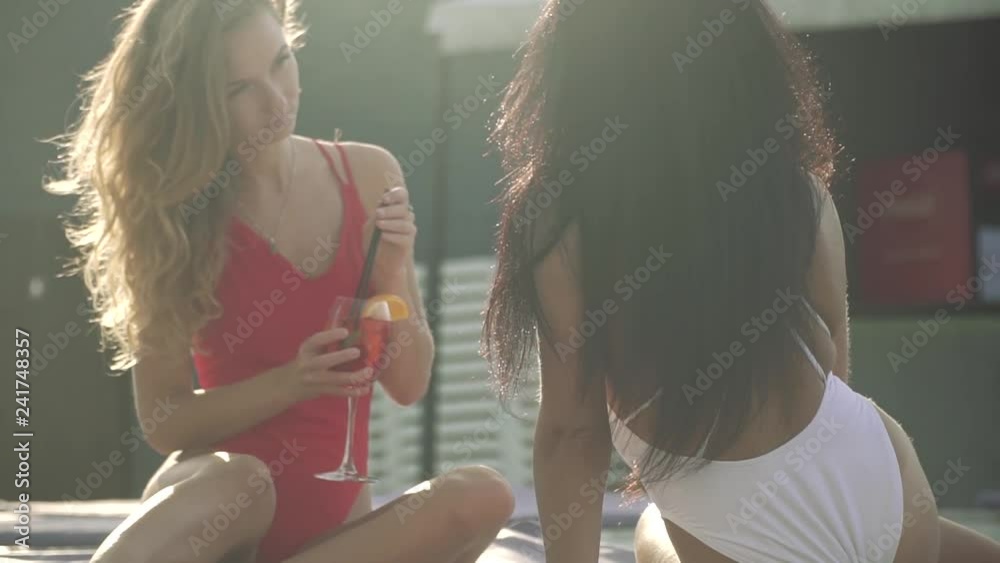 Two sexy girlfriends lying on chaise longue and holding freshly made cocktails.