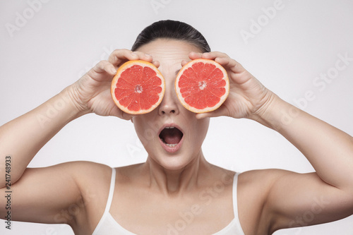 Closeup portrait of suprized woman with perfect skin smiling after cream, balm, mask, lotion, holding half of grapefruit and covering her eye, isolated on grey background. Indoor, studio shot. photo