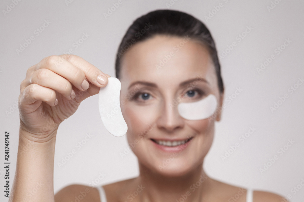 Patch Under Eyes. Beautiful middle aged woman with perfect skin showing white hydrogel patches with lifting anti-wrinkle collagen effect, gray background, indoor, studio shot, copy space, isolated Patch Under Eyes. Beautiful middle aged woman with perfect skin showing white hydrogel patches with lifting anti-wrinkle collagen effect, gray background, indoor, studio shot, copy space, isolated