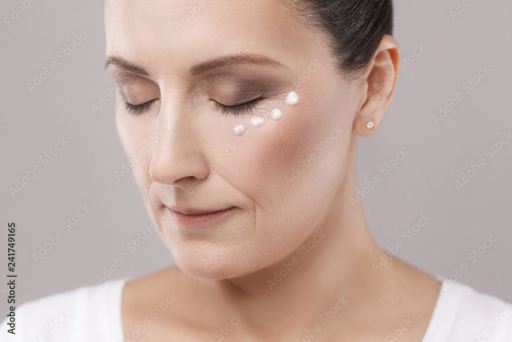 Portrait of middle aged woman with perfect face skin applying cosmetic cream on skin near eyes with closed eyes isolated on grey background. Beauty and skin care concept,copy space,studio shot, indoor Portrait of middle aged woman with perfect face skin applying cosmetic cream on skin near eyes with closed eyes isolated on grey background. Beauty and skin care concept,copy space,studio shot, indoor