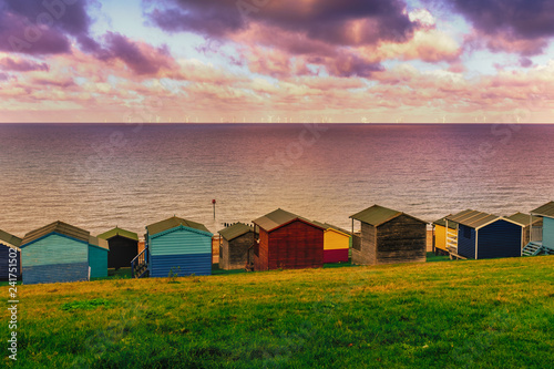Moody cloudy weather creating patches of sunlight between the clouds on the sea in Tankerton, Whitstable in Kent.  Rows of wooden beach huts line the coastline with green sloping banks behind them photo