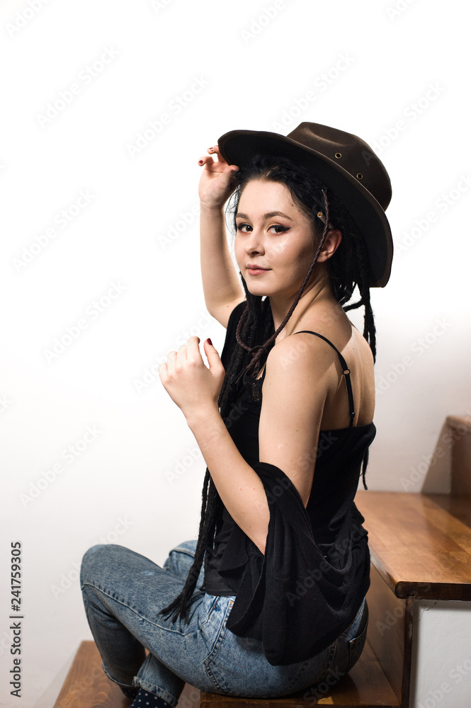 Cute hipster girl wearing a cowboy hat, black top and blue jeans posing on a staircase against a white background. Cute hipster girl wearing a cowboy hat, black top and blue jeans posing on a staircase against a white background.