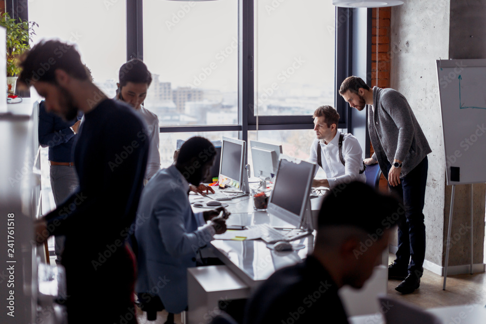 Multi Ethnic group of male architectures and web-designers standing randomly in the big spacious open space office with modern loft interior and computer desks. Routine working process.