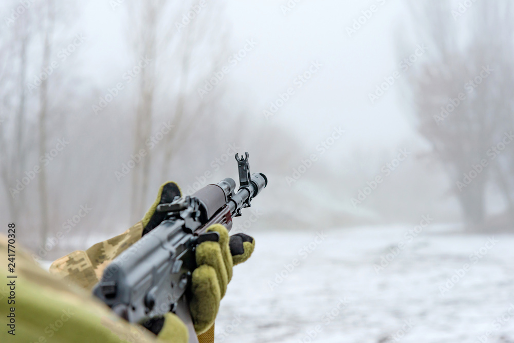 A military soldier takes aim with an automatic rifle and holds it in his mittens in winter.