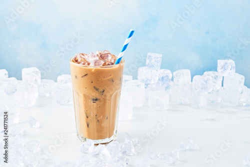 Summer drink ice coffee with cream in a tall glass with straw surrounded by ice on white marble table over blue background. Selective focus, copy space for text.