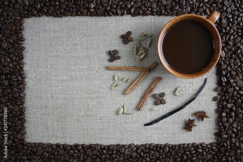 Obramowanie z ziaren kawy, wewnątrz na tkaninie z juty filiżanka czarnej kawy i przyprawy: ziarna kardamonu, laski cynamonu, wanilia i anyż