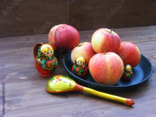 Composition of red and yellow apples on cast iron plate with traditional russian nesting dolls matrioshka and painted spoon on shabby wooden brown background