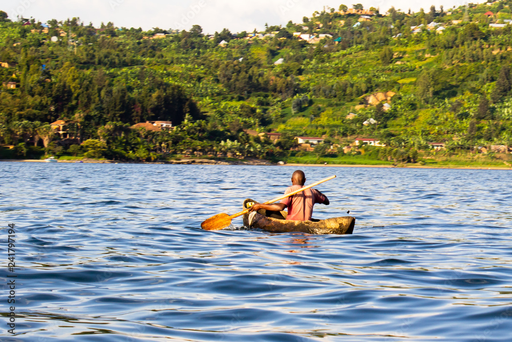 Back view of an African man paddling a dugout canoe in the river, fishing on a bright beautiful day.