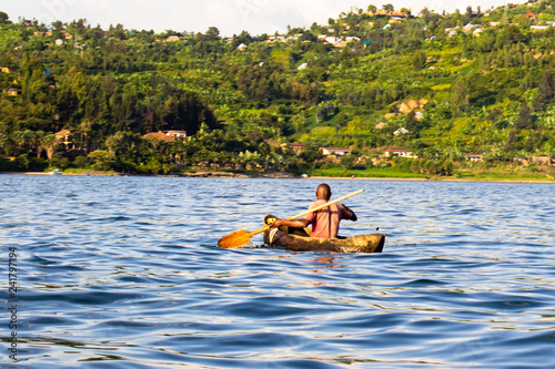 Back view of an African man paddling a dugout canoe in the river, fishing on a bright beautiful day.