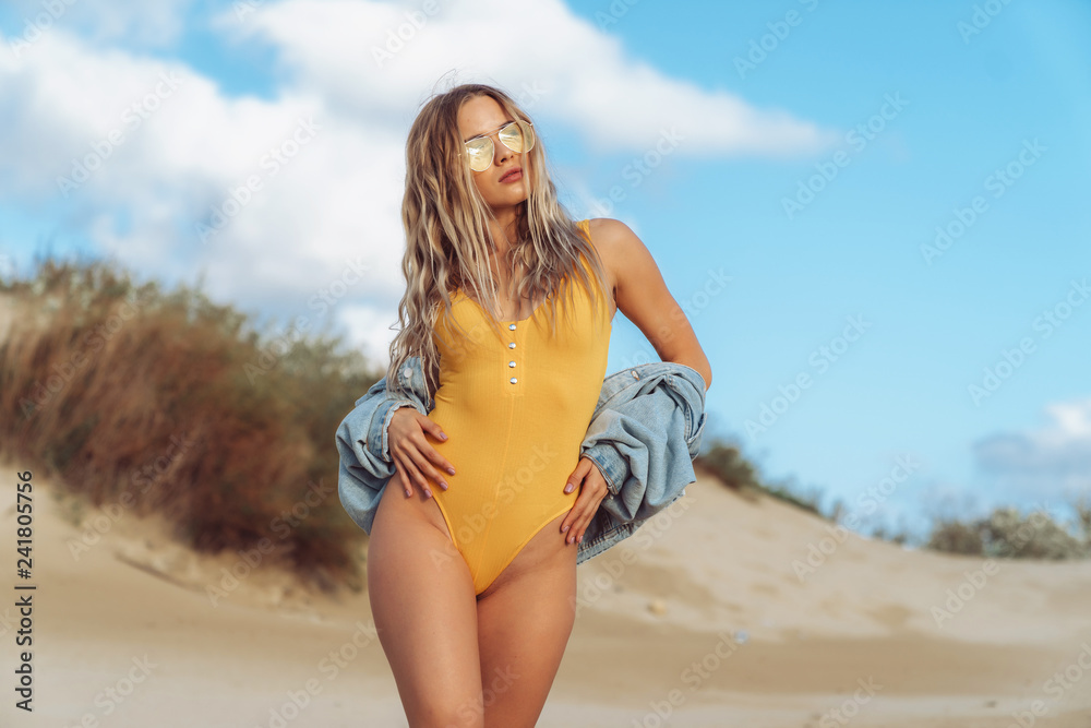 Attractive girl in yellow swimsuit and denim jacket enjoys relaxing on white sand beach