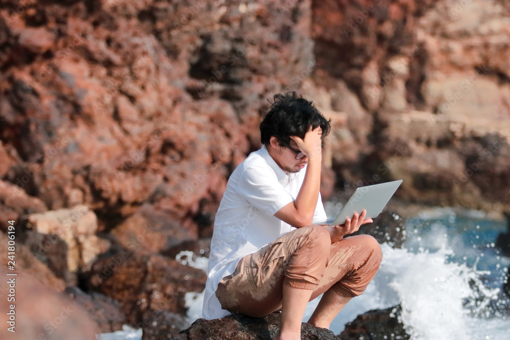 Frustrated anxiety young Asian man with laptop suffering from severe trouble at sea shore.