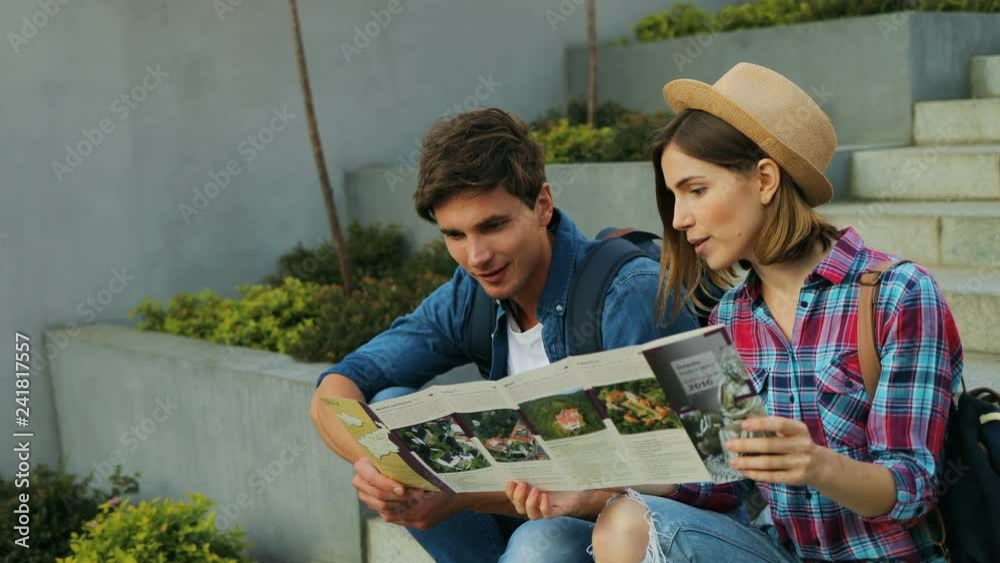 Beautiful young couple is holding a touristic map and smiling while sitting on stairs outdoors.