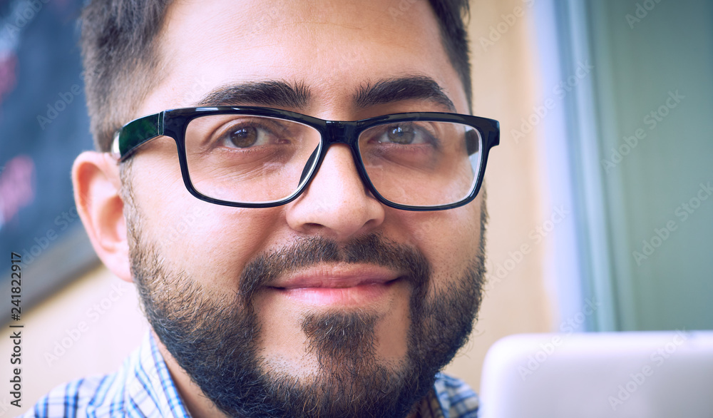 Young bearded businessman in glasses, dressed in plaid shirt, sitting at table in cafe and use smartphone. Man holding smartphone and looking at its screen. Young bearded businessman in glasses, dressed in plaid shirt, sitting at table in cafe and use smartphone. Man holding smartphone and looking at its screen.