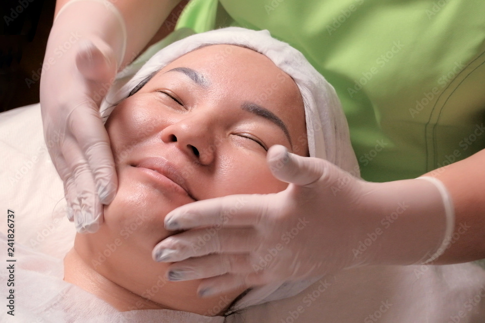 Rejuvenating cosmetological procedure for a Muslim woman. The hands of a beautician in white gloves put the cream around the eyes on the face of an Asian patient. Face massage.