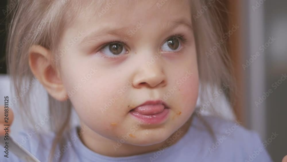 A little girl eats a spoonful of vegetable soup soiling her face and carefully watches TV A little girl eats a spoonful of vegetable soup soiling her face and carefully watches TV
