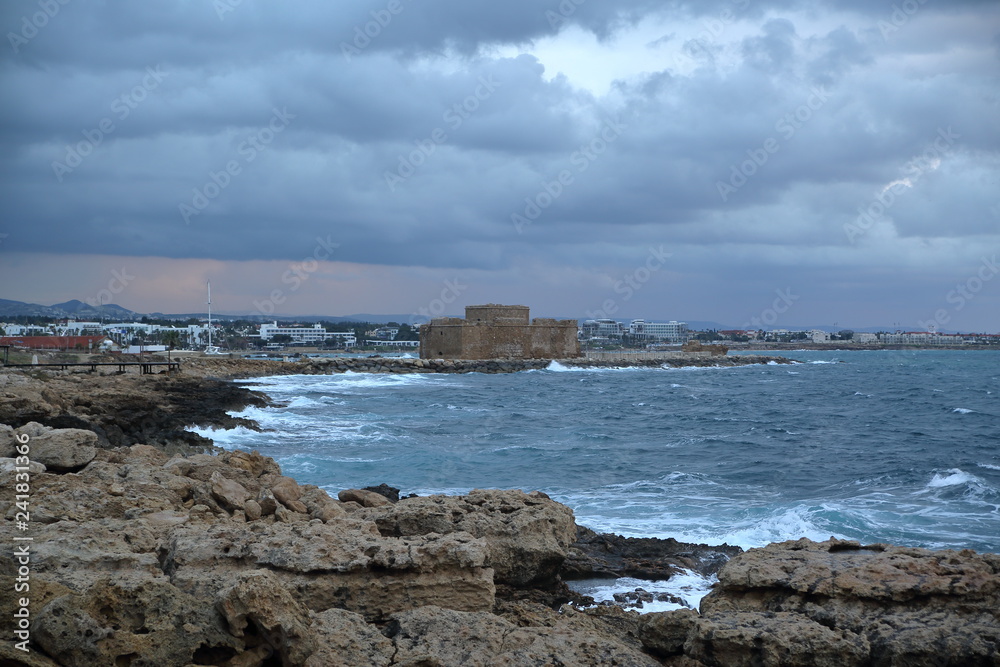 Krajobraz wybrzeża w Pafos, Cypr, skalisty brzeh, lekko wzburzone morze, średniowieczny ufortyfikowany zamek na cyplu, w oddali zabudowa mista, wzgórza