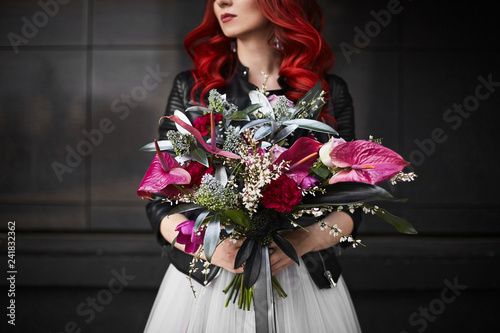 Fashionable and beautiful model girl with red hair and bright makeup, in a white wedding dress and in a leather jacket, with a big and luxury bouquet of exotic flowers in her hands, posing outdoors