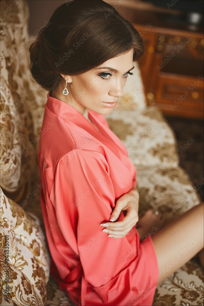 Fashionable and beautiful brunette model girl with perfect sexy body and with blue eyes, in a pink peignoir, sits on vintage sofa and posing at luxury interior