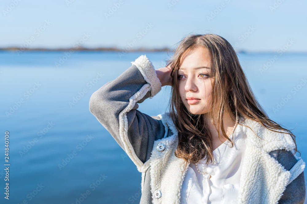 A girl in a light sheepskin coat stands on the bank of the river.