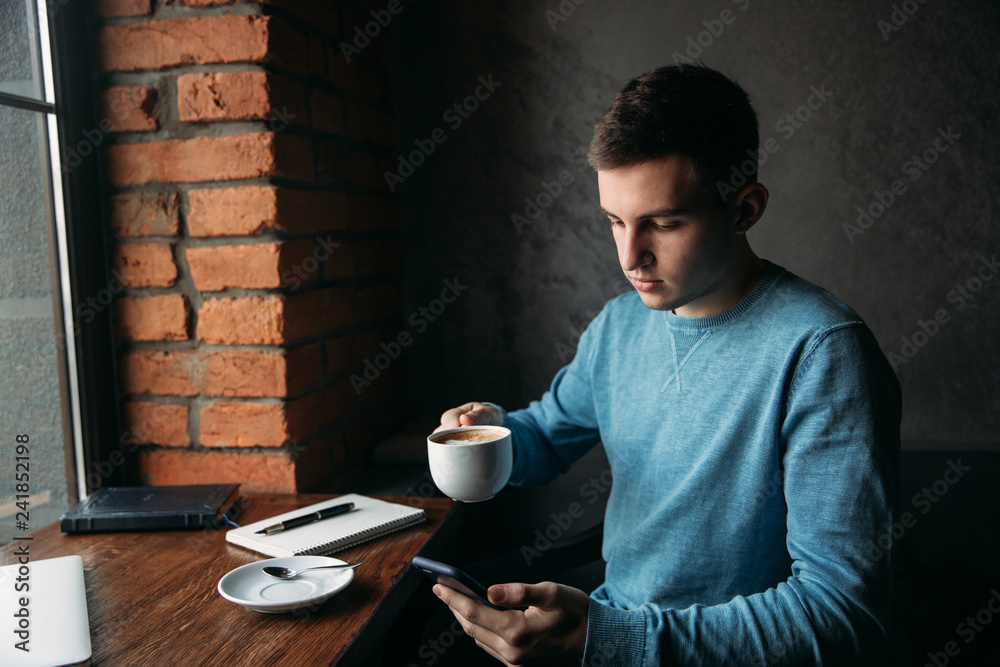 Student using phone inside a cafe while drinking coffee Student using phone inside a cafe while drinking coffee