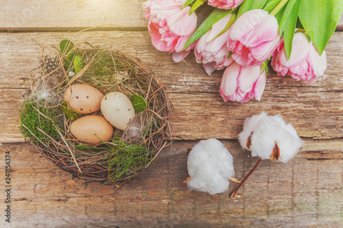 Spring greeting card. Easter eggs in nest with moss and pink fresh tulip flowers bouquet on rustic shabby wooden background. Easter concept. Flat lay top view copy space. Spring flowers tulips