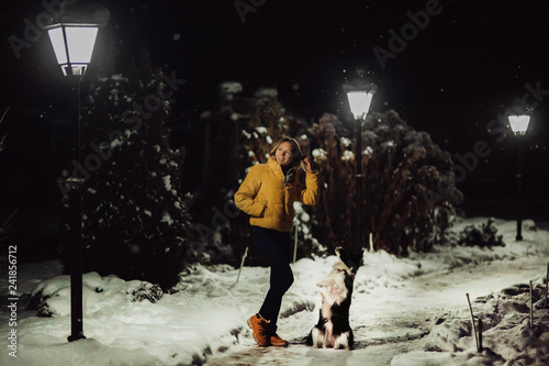 Adorable fluffy cute black and white border colly being trained and petting girl in a park. lights and bushes on the background . Snow is falling