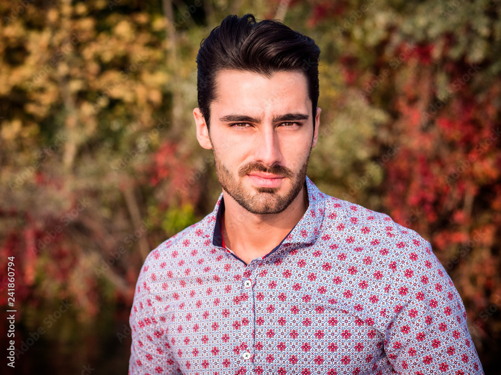 Portrait of contemplative handsome brown haired young man, with short beard, wearing shirt beside picturesque river or lake, looking at camera to a side during fall Portrait of contemplative handsome brown haired young man, with short beard, wearing shirt beside picturesque river or lake, looking at camera to a side during fall