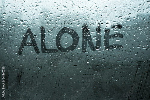 Handwritten word alone on fog up glass window with rain drops in cold dark tones, illustrates sad emotions, unhappy feelings, depression, loneliness and sadness mood