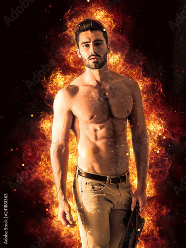 Tattooed muscular man with nude torso with burning fire explosion behind him, on black background