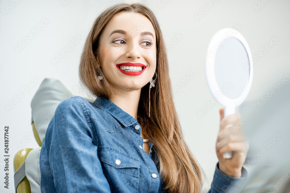 Portrait of a young beautiful woman looking on the mirror enjoying her beautiful smile in the dental office Portrait of a young beautiful woman looking on the mirror enjoying her beautiful smile in the dental office