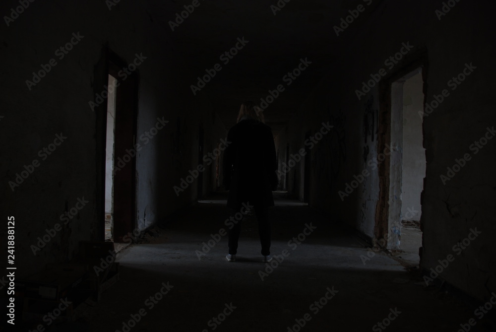 A woman from backside standing in dark corridor of abandoned decay building, urbex 