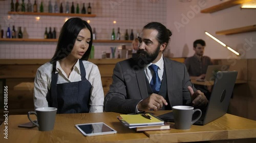 Irritated bearded male manager scolding, criticizing stressed female employee for incompetence at cafe. Sad beautiful employee in apron receiving fair reprimand from restaurant owner indoors. photo