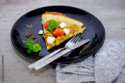 Slice of homemade tart with grilled zucchini, tomatoes and goat's cheese on black plate