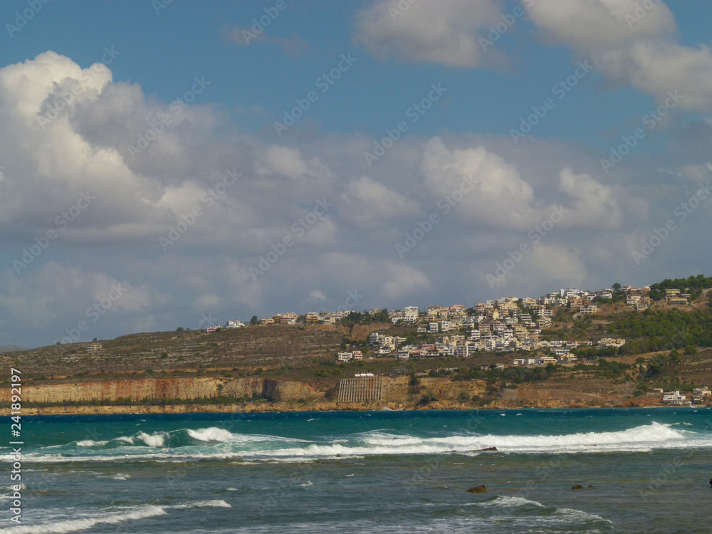 View of the houses of Chania through the strait with the turquoise sea and white lambs, blue sky with white clouds