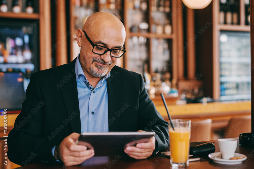 Bearded senior in suit using tablet while sitting in cafeteria. On desk juice and coffee.