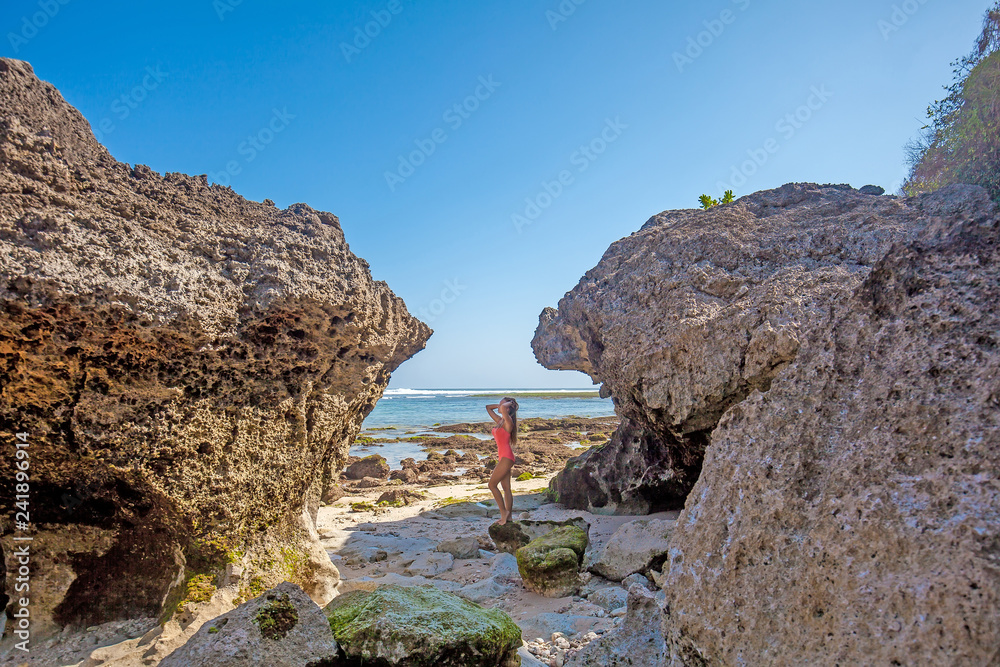 Young sexy woman with long hair dressed in pink swimsuit standing between two huge rocks on the tropical beach near the sea while sky is blue on the Bali island, Indonesia Young sexy woman with long hair dressed in pink swimsuit standing between two huge rocks on the tropical beach near the sea while sky is blue on the Bali island, Indonesia