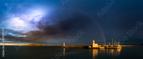 Panorama of Howth Harbour in County Dublin under a dramatic stormy cloudy sky with a rainbow spreading from the Eye of Ireland across to a lighthouse and moored sailing boats bathed in sunlight.