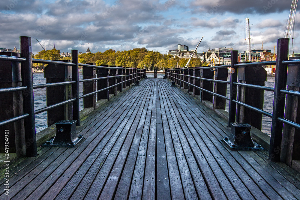 Obraz premium Landscape view of wooden platform on the river Thames.Trees and buildings in the background. London, United Kingdom.