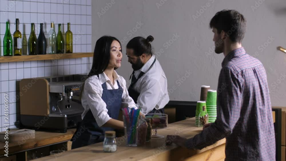 Beautiful mixed race waitress in apron taking order from male customer at coffee shop and stylish barista preparing delicious coffee. Positive friendly cafe owner taking drink order at counter bar.