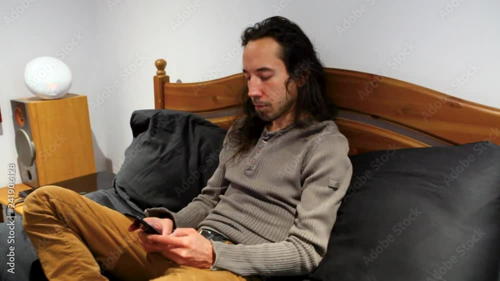 Man on his cellphone in bed. Man with long hair typing on his cellphone, sitting in his bed in bedroom with painting - panning left Man on his cellphone in bed. Man with long hair typing on his cellphone, sitting in his bed in bedroom with painting - panning left