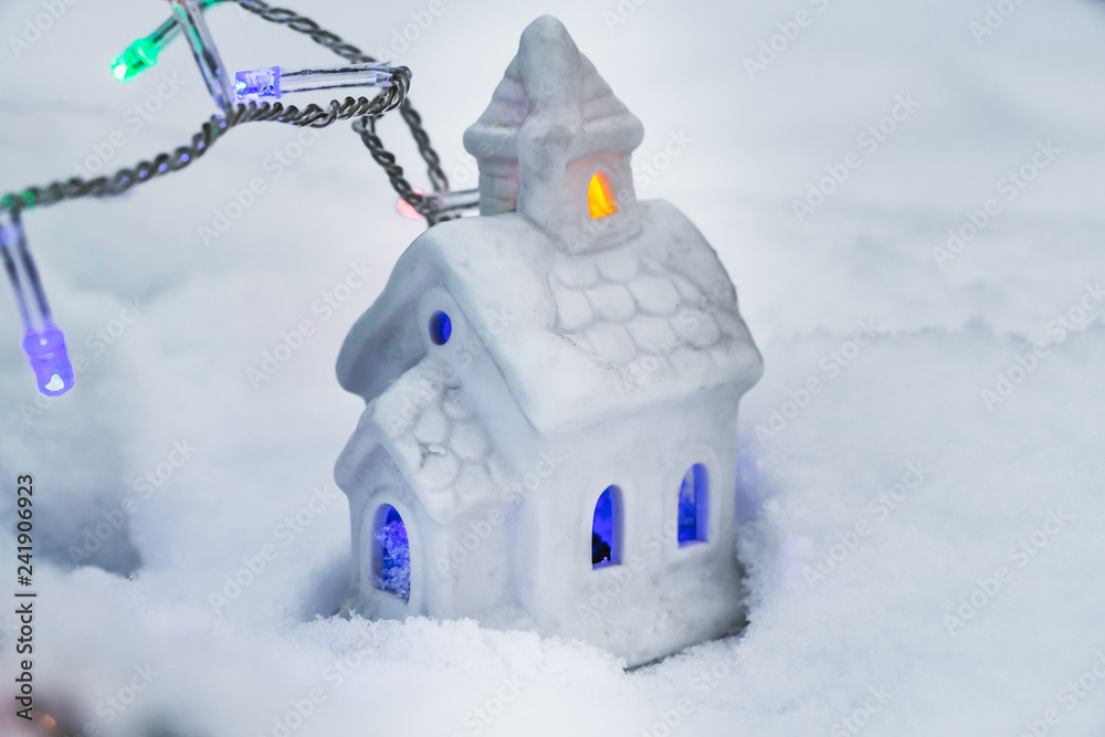 Naklejka premium A beautiful fabulous little white house with yellow and blue lights in the windows and garlands of burning colorful lamps on a white snow background in the holiday evening