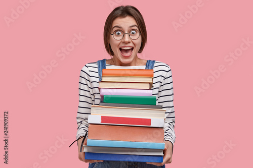Waist up shot of joyful student in round spectacles, holds pile of books, prepares for seminar or writing report, opens mouth widely, isolated over pink background. Intelligent female studies indoor