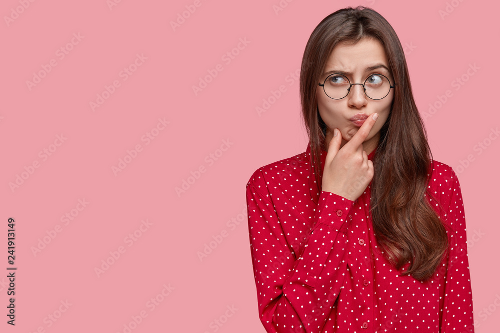 Young attractive woman with thoughtful expression, thinks about question, feels doubt, raises eyebrows, holds chin, dressed in formal clothes, isolated over pink background with free space on left