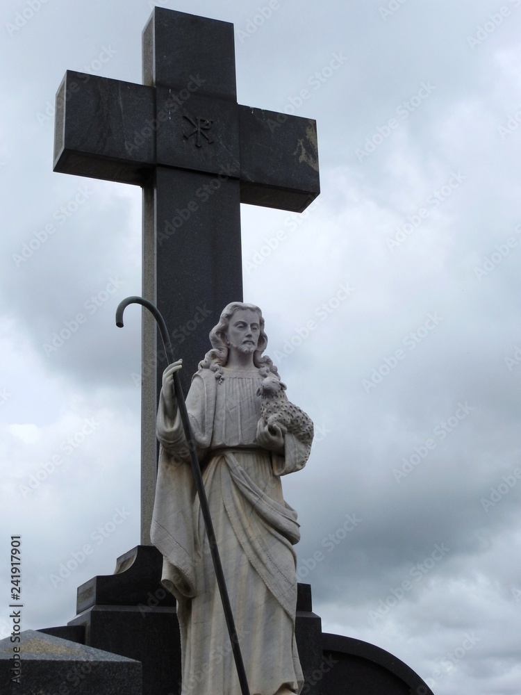 Scene in a graveyard: statue of Jesus, the good shepherd, holding a sheep and a staff. Behind, a large religious cross. Cloudy day.